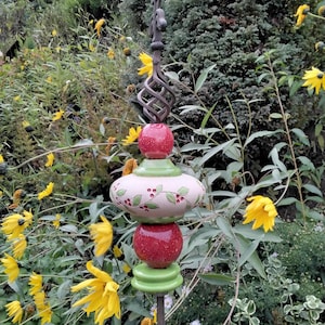 Könnte beinhalten: Ein dekorativer Gartenstecker mit Metallstab und Keramikornamenten. Die Ornamente umfassen rote Kugeln, eine weiße und grüne ovale Form mit Beerenmuster und grüne Sockel. Der Stecker ist von gelben Blumen und Grün umgeben.
