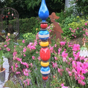 Könnte beinhalten: Eine farbenfrohe Gartenskulptur mit einer blauen tropfenförmigen Spitze. Die Skulptur besteht aus gestapelten, abgerundeten Abschnitten in Rot, Gelb und Blau, inmitten eines Gartens mit rosa Rosen und grünem Laub. Ein dekoratives Element für den Außenbereich.