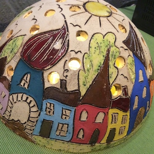 Może przedstawiać: Ceramiczna lampa w kształcie kopuły z ręcznie malowaną sceną wioski. Lampa przedstawia kolorowe domy, drzewa i wzór słońca. Kopuła ma kilka otworów, które emitują ciepłą poświatę po zapaleniu.
