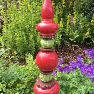 Pode incluir: Uma estaca de jardim decorativa com uma parte superior e inferior de cerâmica vermelha salpicada. A estaca tem elementos de cerâmica verde e castanha no meio. A estaca de jardim está colocada num jardim exuberante com folhagem verde e flores amarelas e roxas.