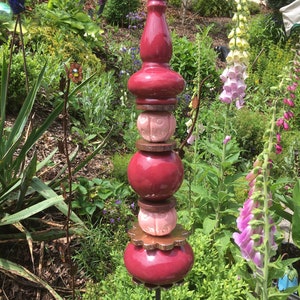 Könnte beinhalten: Ein dekorativer Gartenpfahl mit einem gestapelten Design. Er verfügt über weinrote, abgerundete Formen und ein hellrosa, knollenförmiges Element, alles auf einer dunklen Metallstange. Die Gartenkunst ist von üppigem Grün und bunten Blumen umgeben.