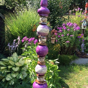 Może przedstawiać: Dekoracyjny palik ogrodowy o warstwowym wzorze. Posiada naprzemienne fioletowe, kuliste i wzorzyste elementy ceramiczne. Szczyt ma kształt bulwiasty, a palik jest osadzony w ogrodzie z zielonymi liśćmi i fioletowymi kwiatami.