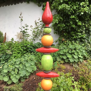 Könnte beinhalten: Ein farbenfroher Gartenstecker mit einer roten tropfenförmigen Spitze, gestapelt mit orangefarbenen und grünen Kugeln, roten Scheiben und strukturierten grünen Kugeln. Die dekorative Außenkunst steht vor einem Hintergrund aus üppigem grünem Laub.
