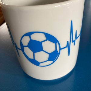 Könnte beinhalten: Weiße Keramiktasse mit einem blauen Fußball und einer blauen Herzschlaggrafik. Die Tasse hat ein weißes Innenleben.