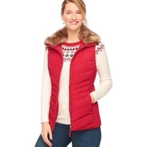 May include: A red quilted vest with a faux fur collar is worn over a cream-colored sweater with a festive pattern. The vest has a full-length zipper and side pockets. The person is wearing blue jeans.