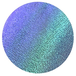 Puede incluir: Muestra circular de maquillaje iridiscente con una superficie texturizada. El color cambia de un azul-púrpura a la izquierda a un verde-turquesa a la derecha. El maquillaje tiene un efecto sutil y brillante.