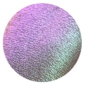Puede incluir: Una muestra circular de maquillaje iridiscente, que transiciona del lavanda al verde azulado y plateado. La superficie tiene una apariencia texturizada, reflejando la luz con un brillo sutil. Este producto es probablemente sombra de ojos o un cosmético similar.