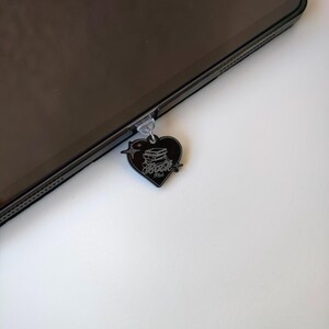 May include: A black heart-shaped charm with the text "Book Hub" and a stack of books, attached to a tablet. The charm is hanging from the tablet's port. The charm has a star and lightning bolt design.