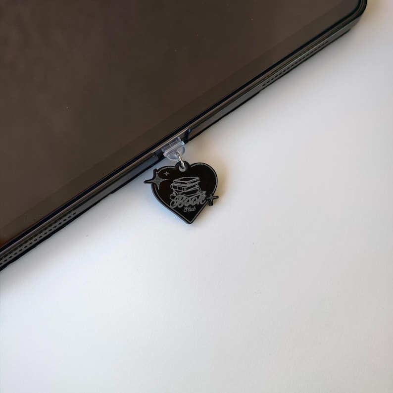 May include: A black heart-shaped charm with the text "Book Hub" and a stack of books, attached to a tablet. The charm is hanging from the tablet's port. The charm has a star and lightning bolt design.