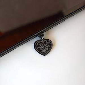 May include: A black heart-shaped charm with the words "Romance Book" and a witch hat design. The charm is attached to a black device. The charm has a decorative border and the Roman numerals "XIXV".