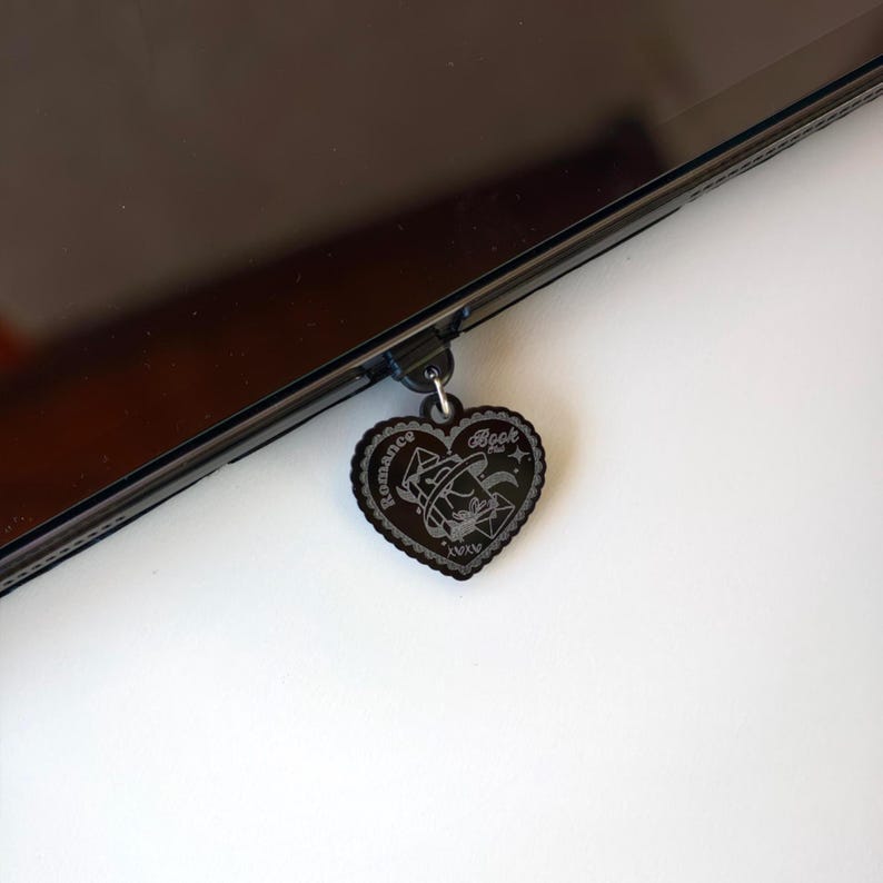 May include: A black heart-shaped charm with the words "Romance Book" and a witch hat design. The charm is attached to a black device. The charm has a decorative border and the Roman numerals "XIXV".