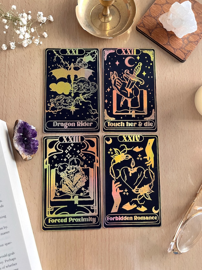 May include: Four tarot cards with black backgrounds and gold foil accents. The cards are numbered XXI, XXII, XXIII, and XXIV. The card titles are "Dragon Rider", "Touch her & die", "Forced Proximity", and "Forbidden Romance".
