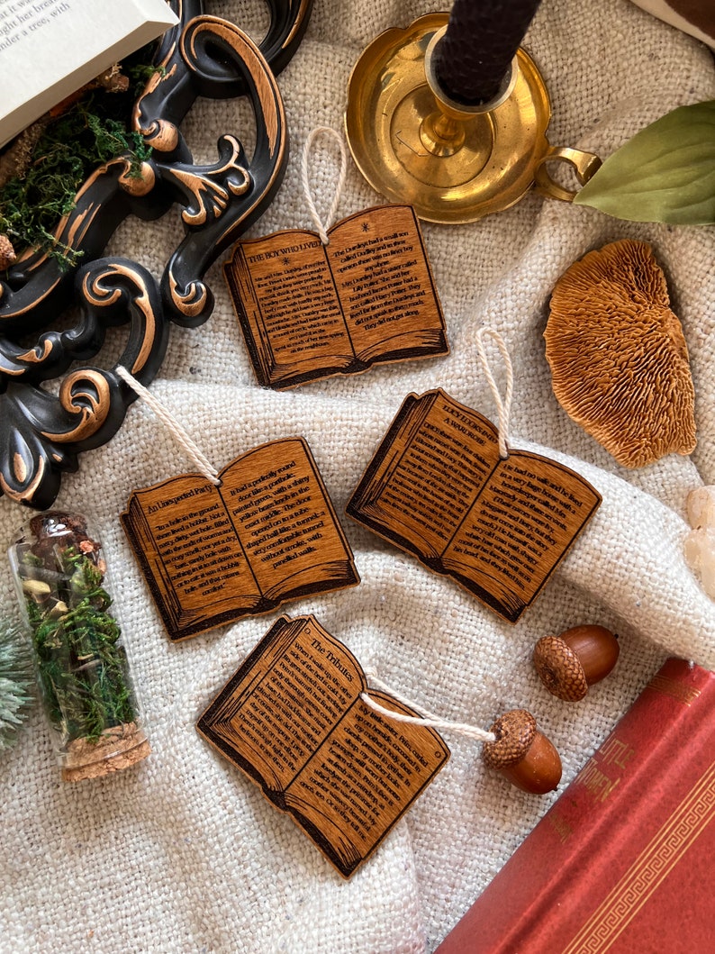 May include: Four wooden book ornaments with text from the Chronicles of Narnia. The ornaments are in the shape of open books with brown wood grain and black text. The titles of the books are "The Boy Who Lived", "The Last Battle", "The Tribute", and "A Wardrobe".