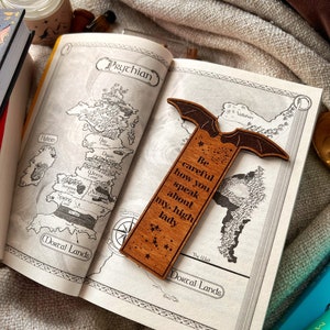 May include: A wooden bookmark with a bat design and the text "Be careful how you speak about my high lady". The bookmark is laying open on a book with a map of a fictional land.