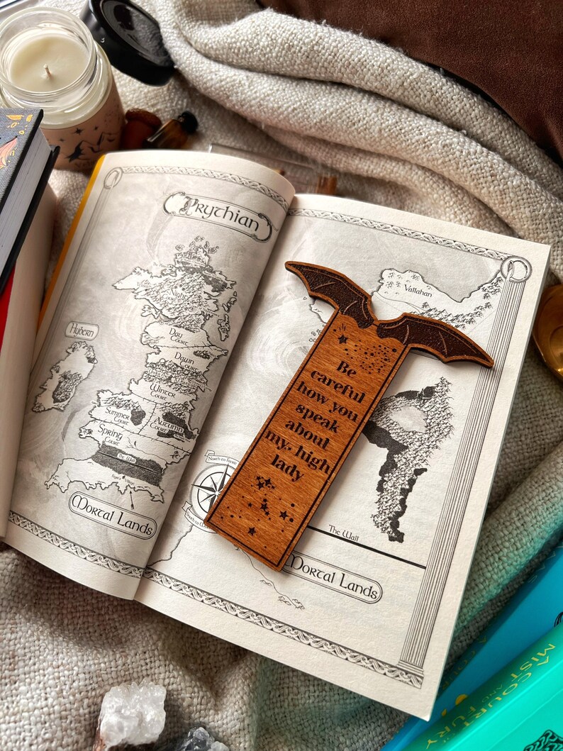 May include: A wooden bookmark with a bat design and the text "Be careful how you speak about my high lady". The bookmark is laying open on a book with a map of a fictional land.