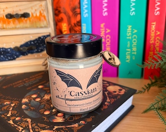 May include: A brown candle jar with a black lid and a white label. The label features a black bat design and the text "Cassian Lord of Bloodshed" and "Sandalwood-Cedar-Balsam Hand Poured Soy". The candle jar is resting on a book with a black cover and a skull design.