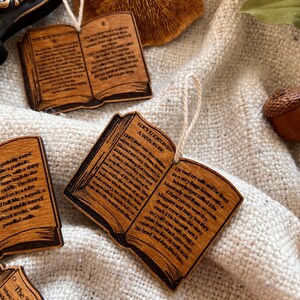 May include: Wooden ornaments shaped like open books with text from popular fantasy novels. The ornaments are decorated with a brown wood grain finish and feature a string for hanging.