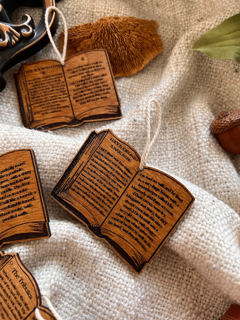 May include: Wooden ornaments shaped like open books with text from popular fantasy novels. The ornaments are decorated with a brown wood grain finish and feature a string for hanging.