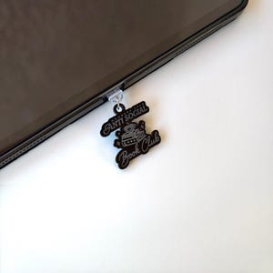 May include: Black phone charm with the words "ANTI SOCIAL" and "Book Club" in a stylized font. The charm is attached to a black tablet. The charm has a small illustration of a stack of books and a crown.