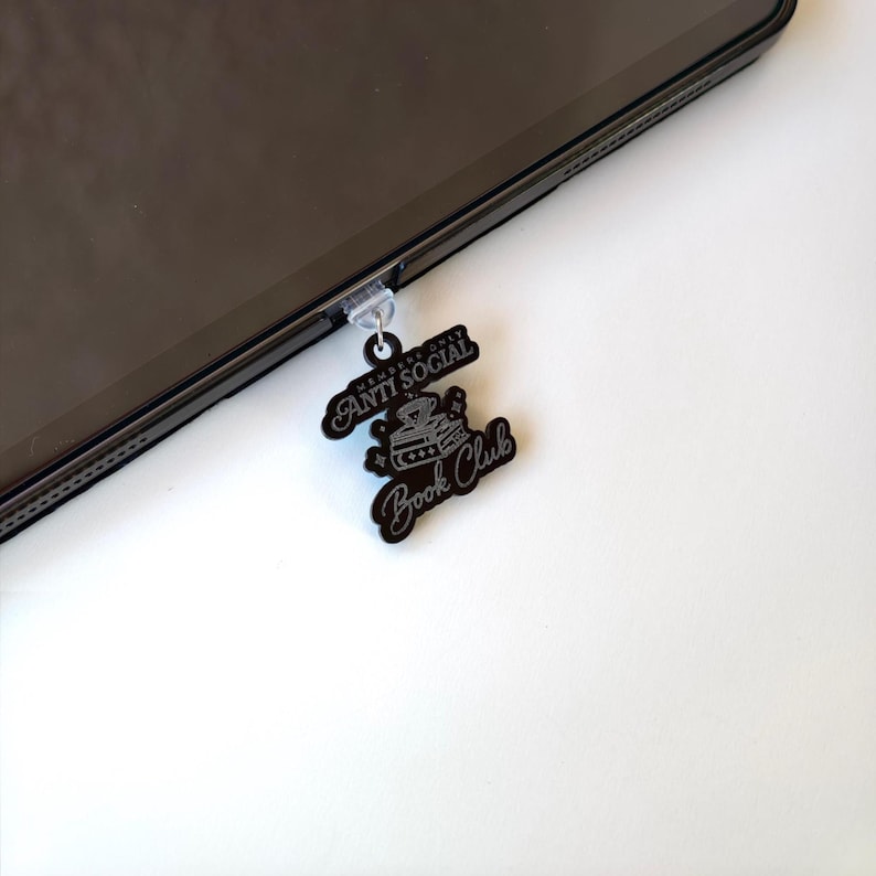 May include: Black phone charm with the words "ANTI SOCIAL" and "Book Club" in a stylized font. The charm is attached to a black tablet. The charm has a small illustration of a stack of books and a crown.