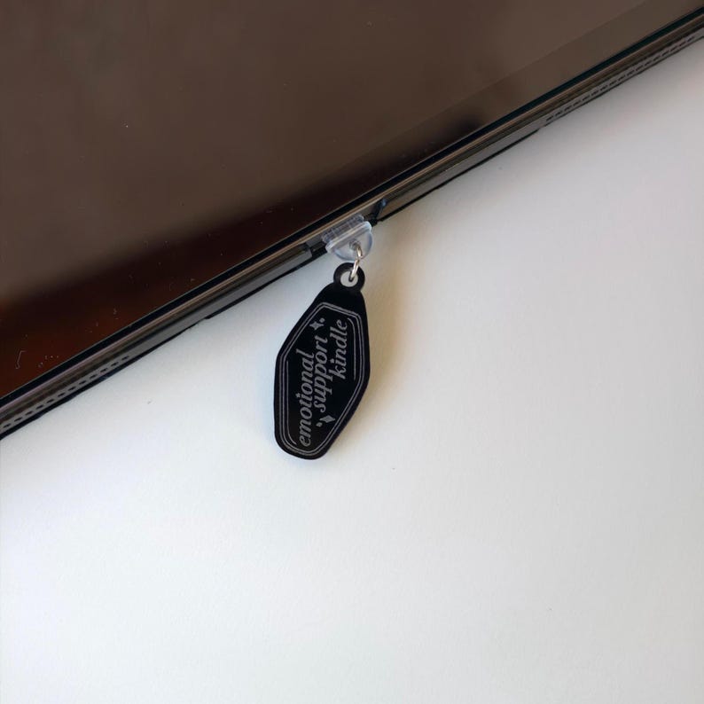 May include: A black acrylic Kindle charm with the words "emotional support Kindle" in white script. The charm is attached to a clear plastic clip, hanging from the edge of a dark electronic device. The background is white.
