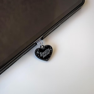 May include: A black heart-shaped charm with the text "Smutty Book Club" and star accents, attached to a tablet. The charm is hanging from a clear plastic piece. The tablet is black and angled on a white surface.