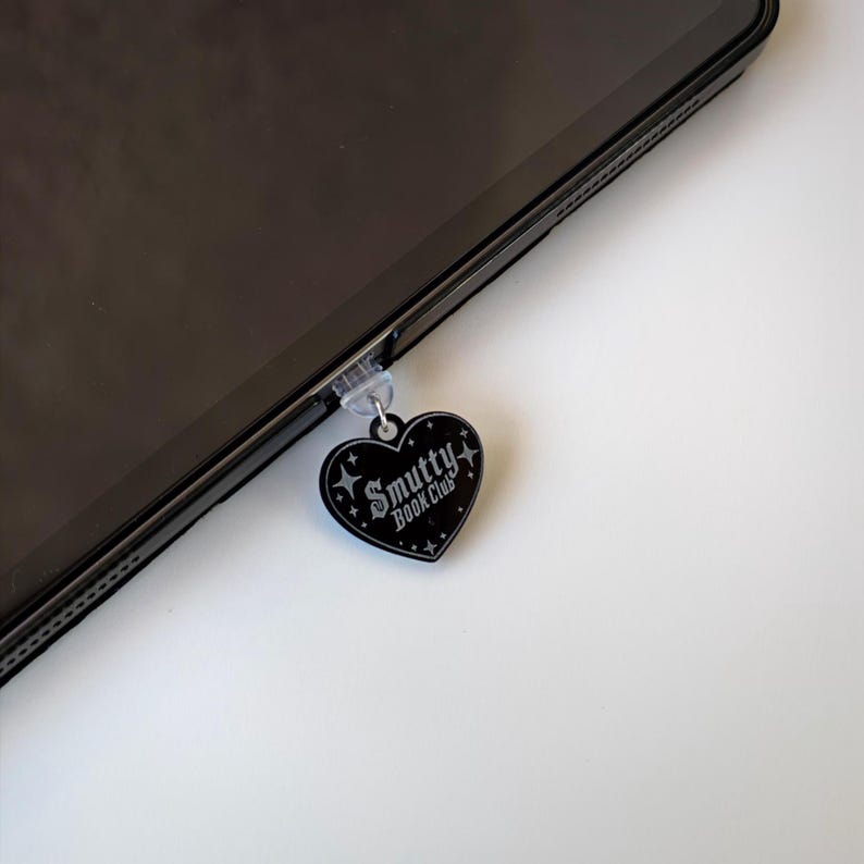 May include: A black heart-shaped charm with the text "Smutty Book Club" and star accents, attached to a tablet. The charm is hanging from a clear plastic piece. The tablet is black and angled on a white surface.