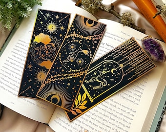 May include: Three gold foil bookmarks with black backgrounds and celestial designs. The bookmarks feature a crescent moon, stars, and a deer, rabbit, and cat. The text on the bookmarks reads "ing Herbs".