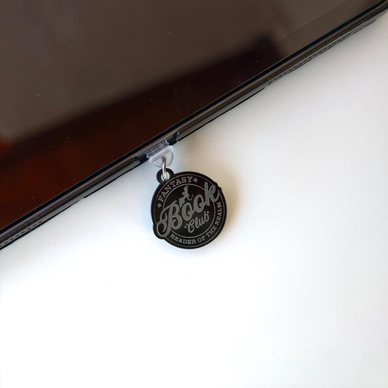 May include: A black and silver Fantasy Book Club charm attached to a tablet. The charm is circular with the words "Fantasy Book Club" and "Reader of the Realm" in a decorative font. The tablet is black and has a clear screen protector.