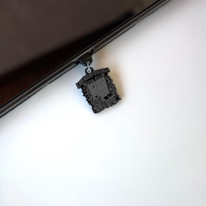 May include: Black metal charm with the text "LATE NIGHT READERS BOOK CLUB MEETS AT MIDNIGHT" and a design of a book, moon, and stars. The charm is attached to a black device.