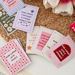 May include: A collection of rectangular reading challenge cards featuring various pastel backgrounds and patterns. Each card displays text prompts such as Read a book with an office romance, Read a book with rivals, and Read a book with a sports romance.