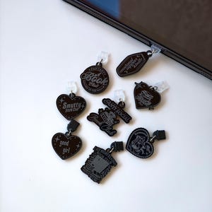 May include: A collection of black acrylic charms with various book-themed designs and text, including "Fantasy Book Club," "Smutty Book Club," and "Anti Social." Each charm has a small clip for attachment.