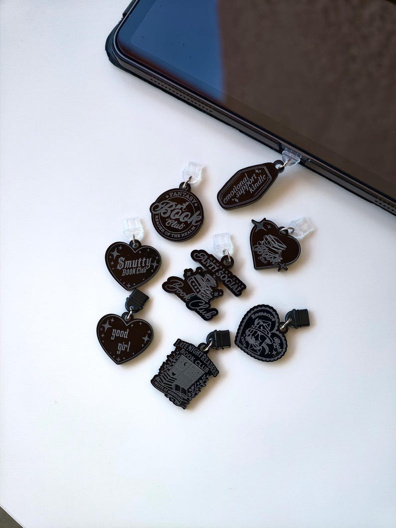 May include: A collection of black acrylic charms with various book-themed designs and text, including "Fantasy Book Club," "Smutty Book Club," and "Anti Social." Each charm has a small clip for attachment.