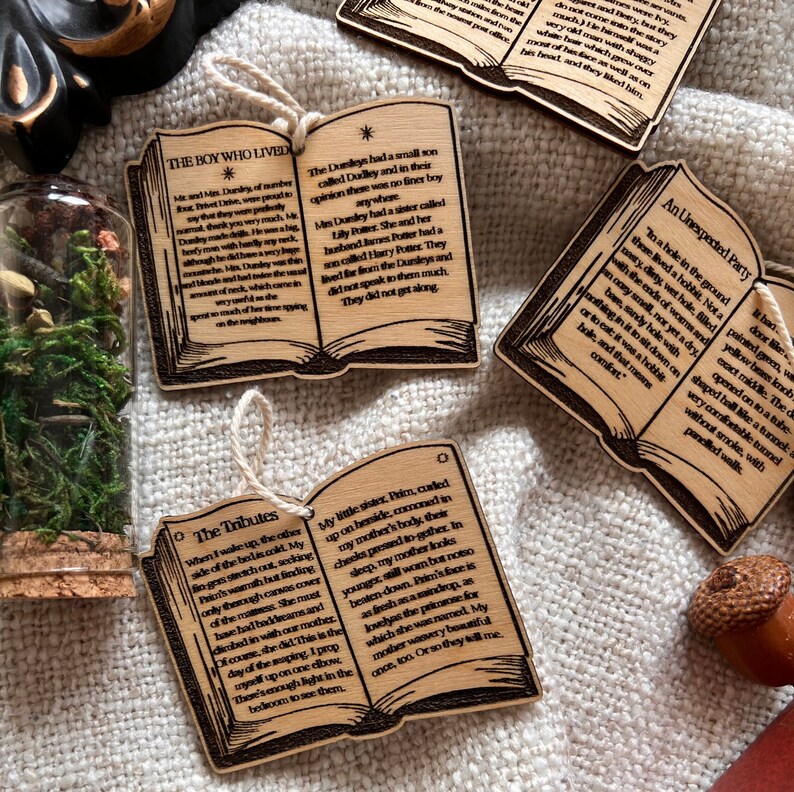 May include: A set of three wooden book ornaments with engraved text from the Harry Potter series. The ornaments are shaped like open books with brown covers and white text. The text on the ornaments reads "The Boy Who Lived", "The Tributes", and "An Unexpected Party".