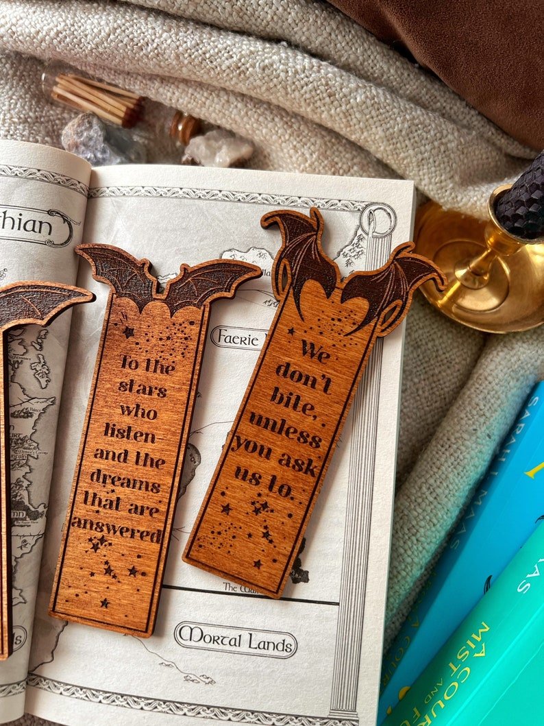 May include: Two wooden bookmarks with a bat design. The bookmarks have a quote about stars and dreams on one and a quote about biting on the other. The bookmarks are on a map with the words "Mortal Lands" printed on it.