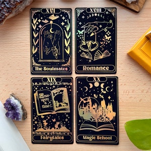 May include: Four tarot cards with gold foil accents on a black background. The cards are numbered XVI, XVIII, XIX, and XX. The card titles are "The Soulmates", "Romance", "Fairytales", and "Magic School".