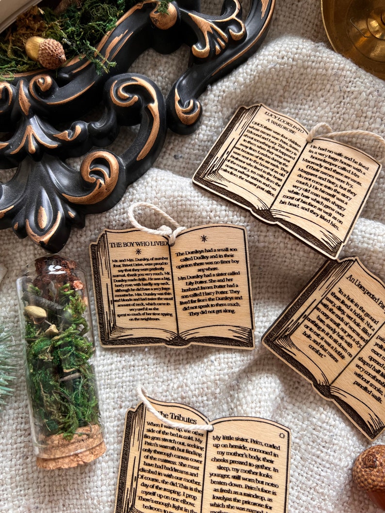 May include: Four wooden book-shaped ornaments with text from the Harry Potter series. The ornaments are decorated with a star and a small illustration of a book. The text on the ornaments includes "The Boy Who Lived", "Lucy Looks Into A Wardrobe", "The Tributes", and "An Unexpected Party".