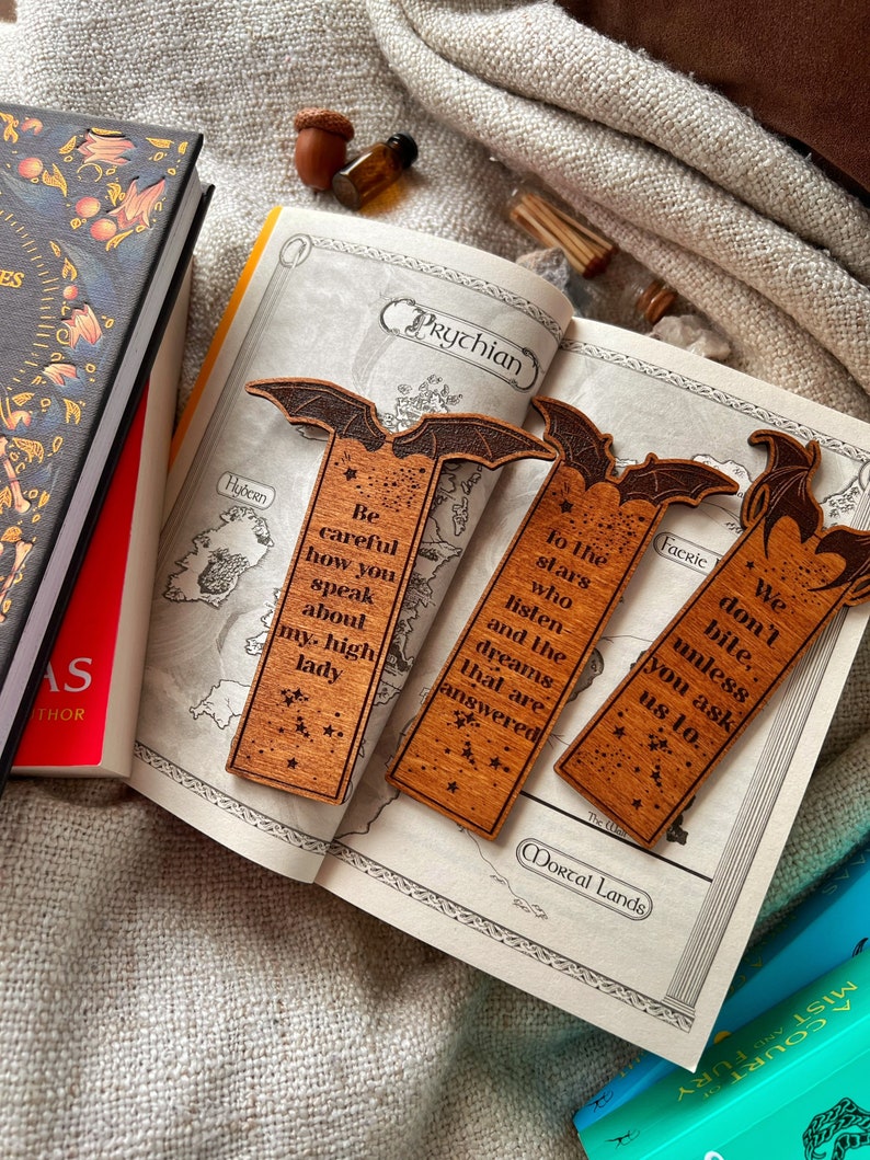 May include: Three wooden bookmarks shaped like bats with different quotes on them. The quotes are "Be careful how you speak about my high lady", "To the stars who listen and the dreams that are answered", and "We don't bite, unless you ask us to."