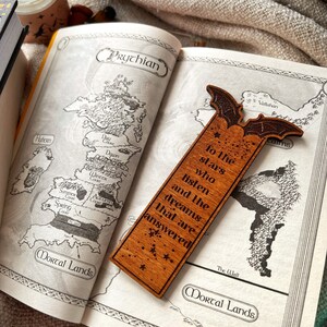 May include: A wooden bookmark with a bat design and the text "To the stars who listen and the dreams that are answered". The bookmark is laying on a book with a map of a fictional land.