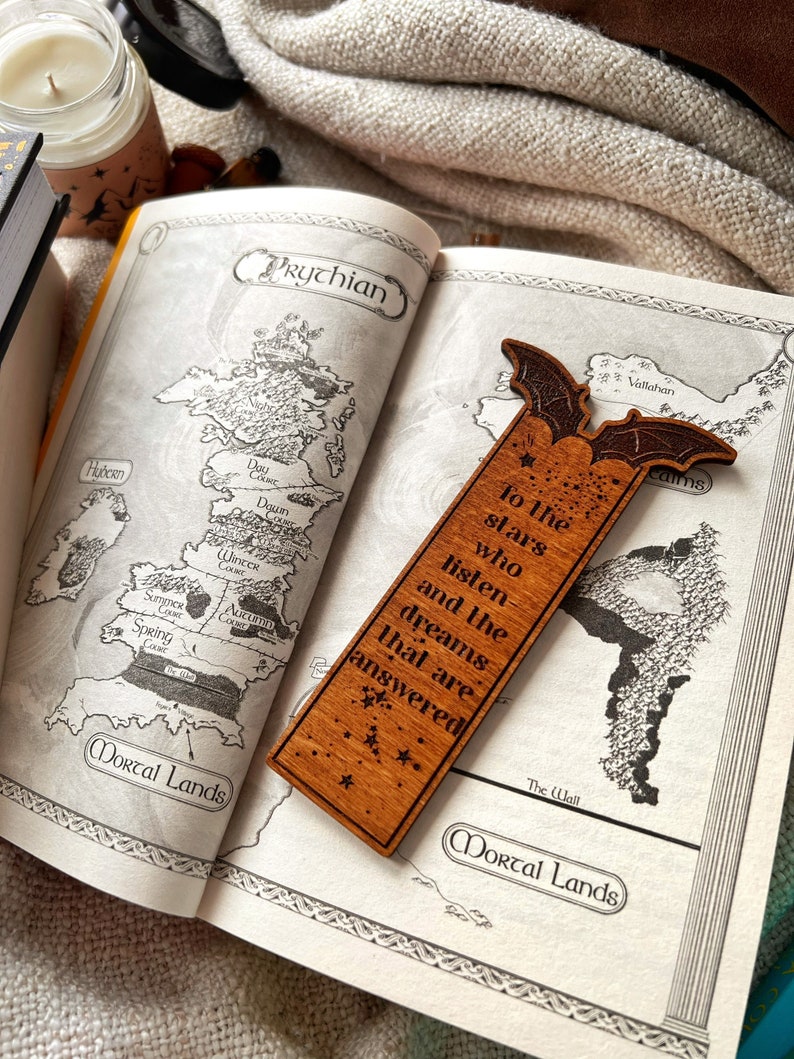 May include: A wooden bookmark with a bat design and the text "To the stars who listen and the dreams that are answered". The bookmark is laying on a book with a map of a fictional land.
