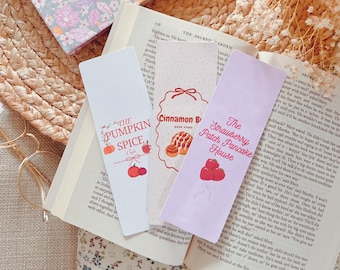 Könnte beinhalten: Drei farbige Lesezeichen mit Illustrationen von Essen und Text. Das erste Lesezeichen ist weiß mit einem Kürbisgewürz-Latte und dem Text "The Pumpkin Spice Cafe". Das zweite Lesezeichen ist rosa mit einem Zimtschnecken und dem Text "Cinnamon Bun Book Store". Das dritte Lesezeichen ist lila mit einer Erdbeere und dem Text "The Strawberry Patch Pancake House".
