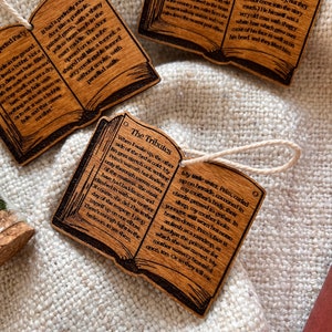 May include: Three wooden book-shaped ornaments with text about hobbits, tributes, and a family living in a large house. The ornaments are on a white fabric background.