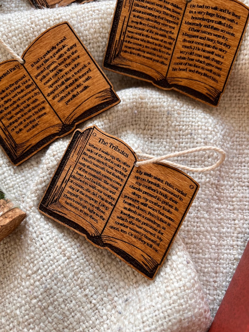 May include: Three wooden book-shaped ornaments with text about hobbits, tributes, and a family living in a large house. The ornaments are on a white fabric background.