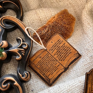 May include: A wooden book-shaped ornament with a brown stain and a white string. The ornament has the text "The Boy Who Lived" and a quote from the Harry Potter series.
