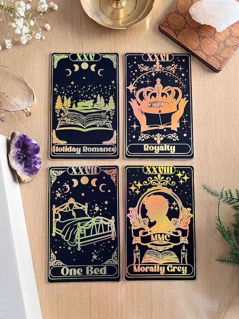 May include: Four tarot cards with gold foil accents on a black background. The cards are numbered XXV, XXVI, XXVII, and XXVIII. The card titles are "Holiday Romance", "Royalty", "One Bed", and "Morally Grey".