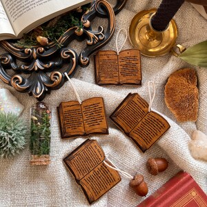 May include: Four wooden ornaments shaped like open books with text printed on them. The ornaments are hanging from twine and are on a white fabric surface. The text on the ornaments includes "The Somnoulous", "The Tribuls", "An Unexpected Party", and "A Day With The Birds".