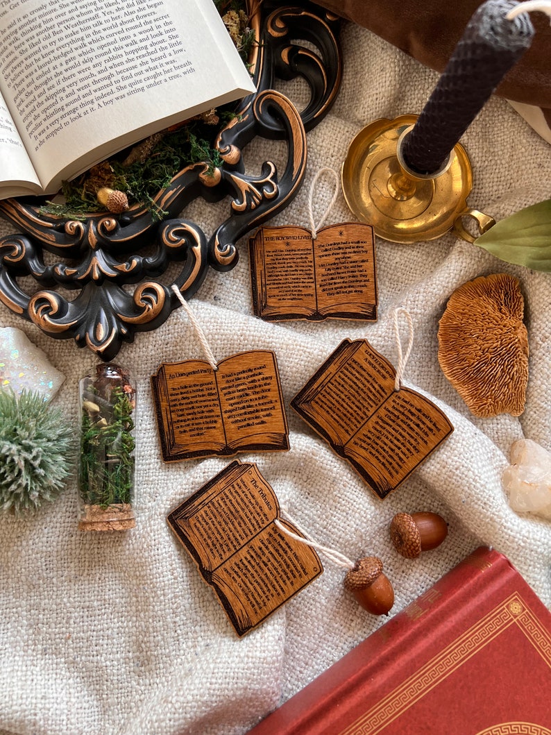 May include: Four wooden ornaments shaped like open books with text printed on them. The ornaments are hanging from twine and are on a white fabric surface. The text on the ornaments includes "The Somnoulous", "The Tribuls", "An Unexpected Party", and "A Day With The Birds".