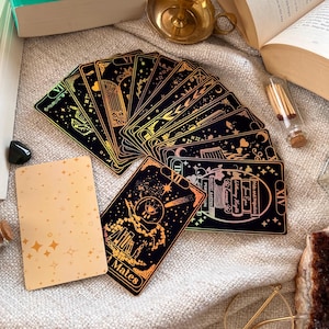 May include: A set of tarot cards with gold foil accents on a black background. The cards are fanned out and show various images, including a castle, a moon, and a star. The cards are on a white fuzzy surface with a gold candle holder, a book, and a small glass jar with matches.