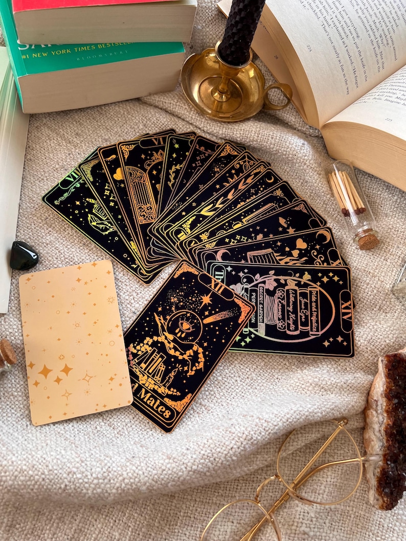 May include: A set of tarot cards with gold foil accents on a black background. The cards are fanned out and show various images, including a castle, a moon, and a star. The cards are on a white fuzzy surface with a gold candle holder, a book, and a small glass jar with matches.