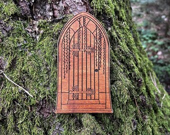 May include: A wooden fairy door with an arched top and detailed etched design. The door has vertical bars, decorative swirls, and hanging vines. It is attached to a moss-covered tree trunk, creating a whimsical, natural scene.